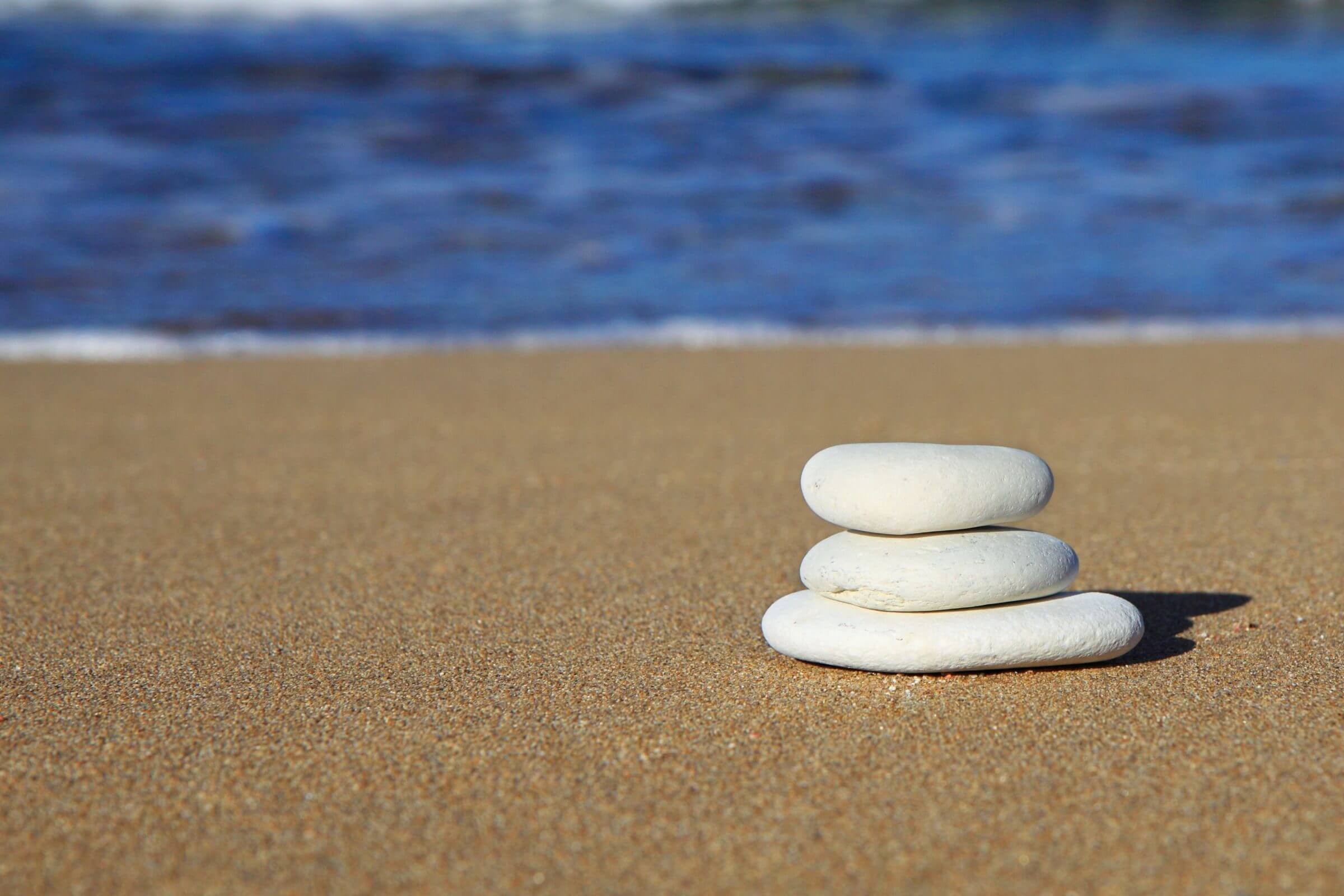 Three pale, flat shaped stones piled on top of one another. In the background is a blurred sea shore.